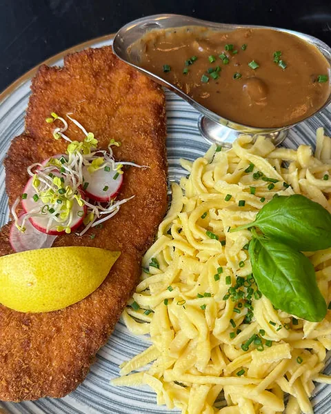 Paniertes Schnitzel mit Spätzle, Bratensoße in der Sauciere und Zitrone auf blau geringeltem Teller
